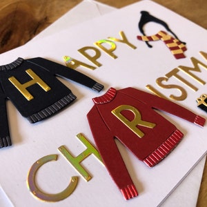 May include: A handmade Christmas card with a festive theme. The card features gold foil lettering that reads "Happy Christmas" and includes cut-out sweater designs with the letters "H" and "R". A penguin and gift box are also included.