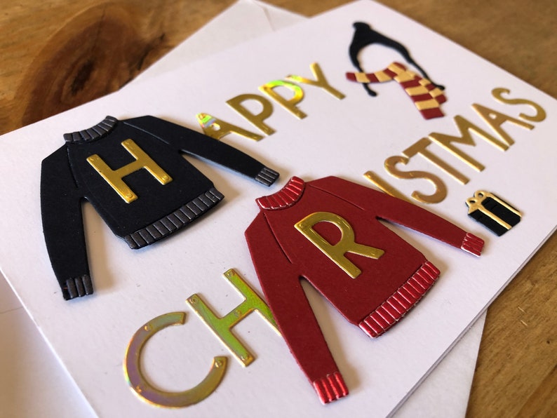 May include: A handmade Christmas card with a festive theme. The card features gold foil lettering that reads "Happy Christmas" and includes cut-out sweater designs with the letters "H" and "R". A penguin and gift box are also included.