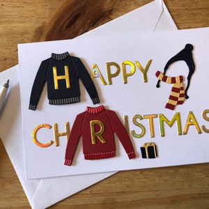 May include: A white greeting card with the words "HAPPY CHRISTMAS" in gold lettering. The card features cut-out sweater designs, a hat, scarf, and a gift box. The sweaters have the letters "H" and "R" on them.