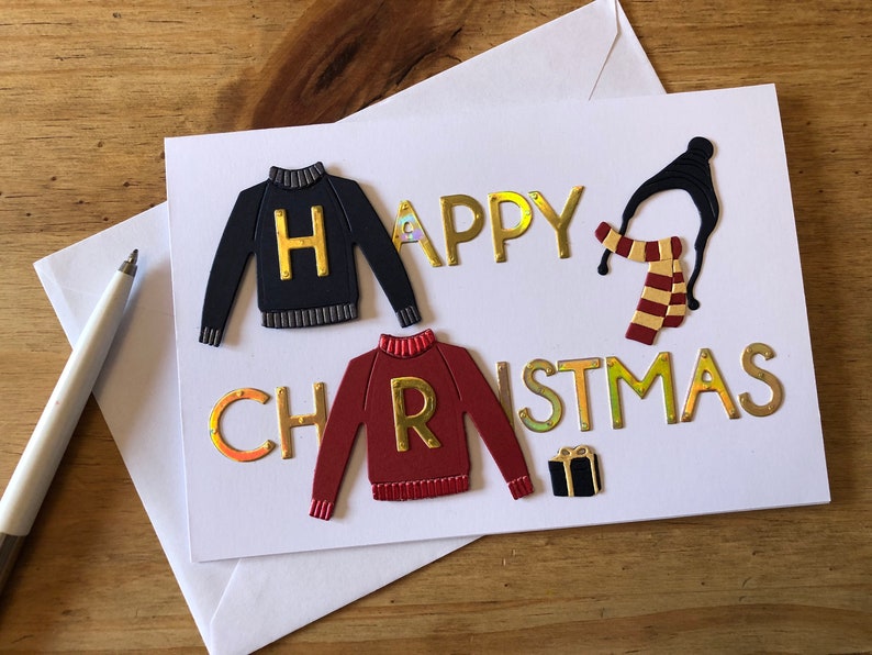 May include: A white greeting card with the words "HAPPY CHRISTMAS" in gold lettering. The card features cut-out sweater designs, a hat, scarf, and a gift box. The sweaters have the letters "H" and "R" on them.