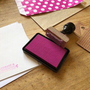 May include: A wooden stamp with text is positioned over a magenta ink pad. A white card with the text "MANDRAKE & MUSHROOMS" is visible. A pink polka dot mailer and brown paper envelope are also in the image.