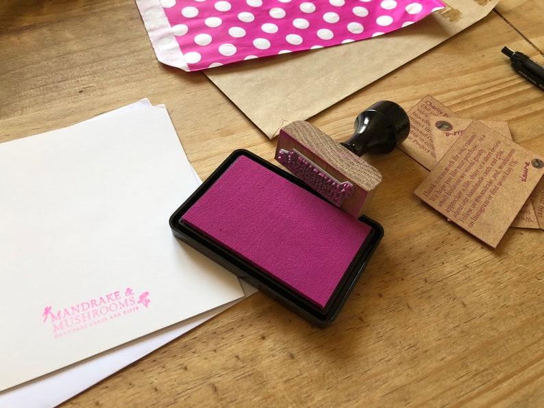 May include: A wooden stamp with text is positioned over a magenta ink pad. A white card with the text "MANDRAKE & MUSHROOMS" is visible. A pink polka dot mailer and brown paper envelope are also in the image.