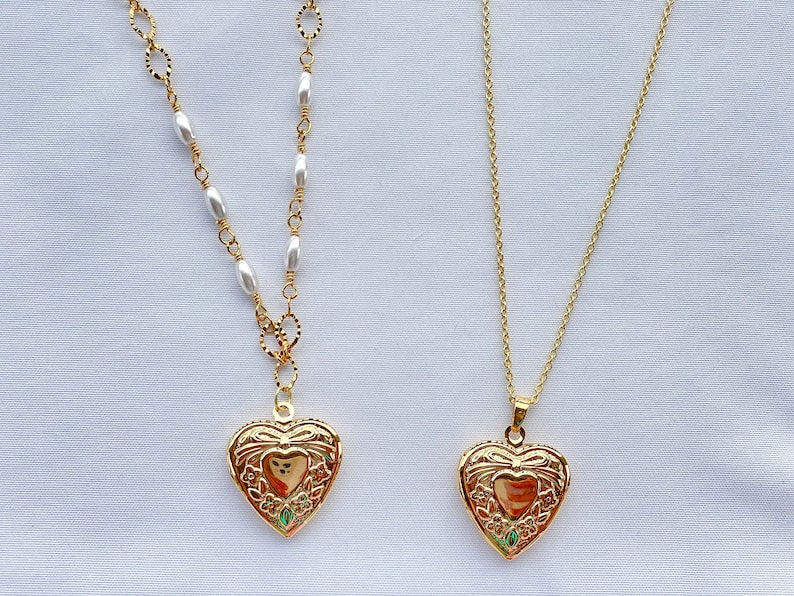 May include: Two gold heart-shaped locket necklaces. One necklace has a chain with white oval beads, the other has a simple gold chain.