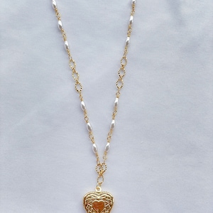 May include: A gold heart-shaped locket pendant hangs from a gold chain with white pearl beads. The locket has an intricate floral design.