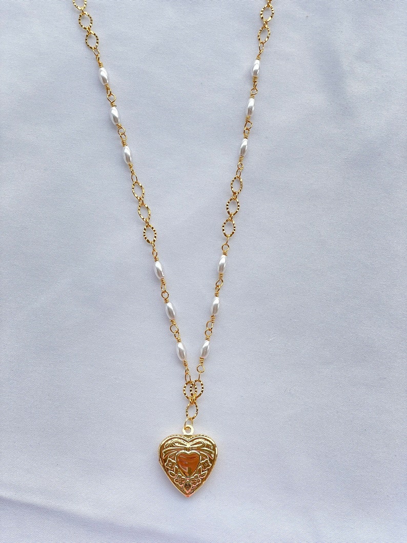 May include: A gold heart-shaped locket pendant hangs from a gold chain with white pearl beads. The locket has an intricate floral design.