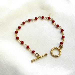 May include: A delicate bracelet featuring small, round red beads strung on a gold-colored chain. The bracelet has a gold-colored toggle clasp. The jewelry is displayed on a white, silky surface.