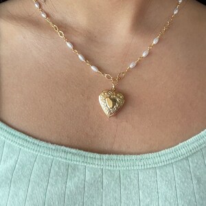 May include: A gold heart-shaped locket necklace with a delicate pearl chain. The locket has an intricate design and is worn on a person's neck.