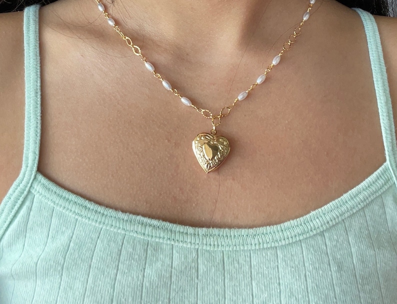 May include: A gold heart-shaped locket necklace with a delicate pearl chain. The locket has an intricate design and is worn on a person's neck.