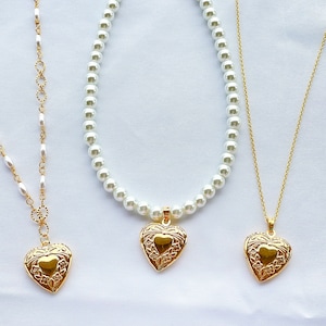 May include: Three gold necklaces with heart-shaped pendants. The necklaces are on a white background. The pendant on the left is a simple heart, the pendant in the middle is a heart with a floral design, and the pendant on the right is a heart with a floral design.