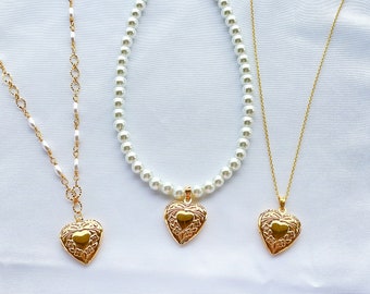 May include: Three gold necklaces with heart-shaped pendants. The necklaces are on a white background. The pendant on the left is a simple heart, the pendant in the middle is a heart with a floral design, and the pendant on the right is a heart with a floral design.