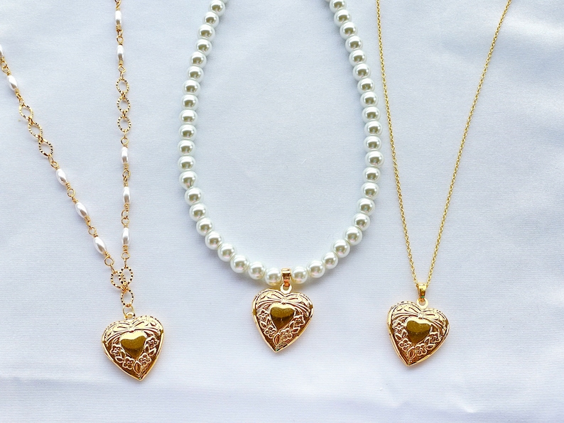 May include: Three gold necklaces with heart-shaped pendants. The necklaces are on a white background. The pendant on the left is a simple heart, the pendant in the middle is a heart with a floral design, and the pendant on the right is a heart with a floral design.