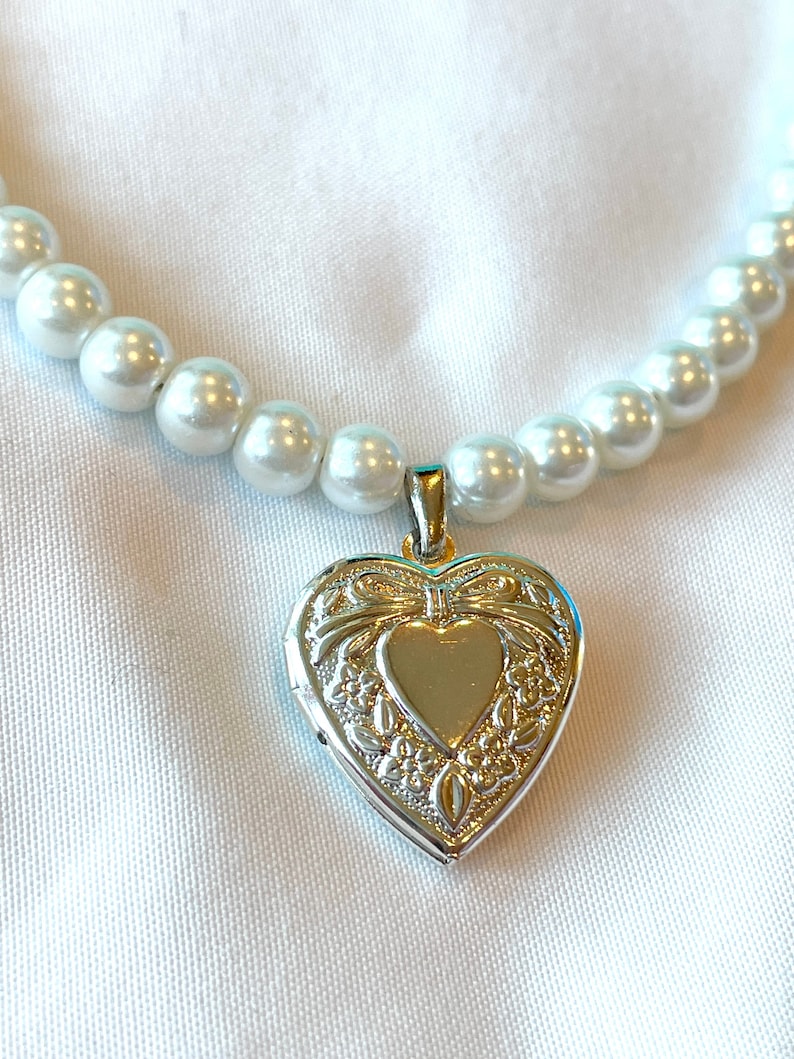 Peut inclure: Un collier de perles blanches avec un pendentif en forme de c&oelig;ur argent&eacute;. Le m&eacute;daillon a un motif floral d&eacute;taill&eacute; et un petit c&oelig;ur au centre.