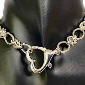 May include: A silver chain necklace with a heart-shaped clasp. The chain is made of interlocking rings.