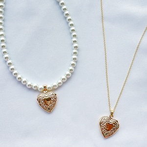 May include: Two necklaces with heart-shaped pendants. The top necklace is a pearl strand with a gold heart pendant. The bottom necklace is a gold chain with a gold heart pendant.