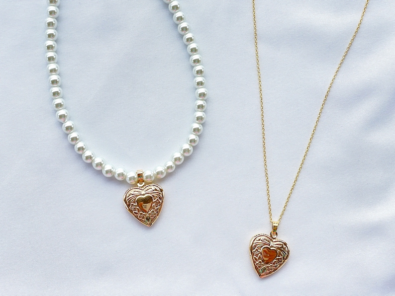 May include: Two necklaces with heart-shaped pendants. The top necklace is a pearl strand with a gold heart pendant. The bottom necklace is a gold chain with a gold heart pendant.