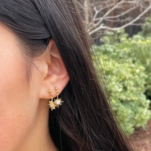 May include: A pair of gold starburst earrings with a single clear rhinestone in the center of each star. The earrings are dangling from a small gold ball.