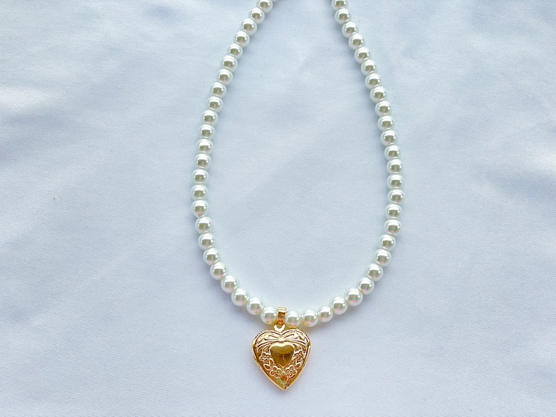May include: A white pearl necklace with a gold heart-shaped locket pendant.