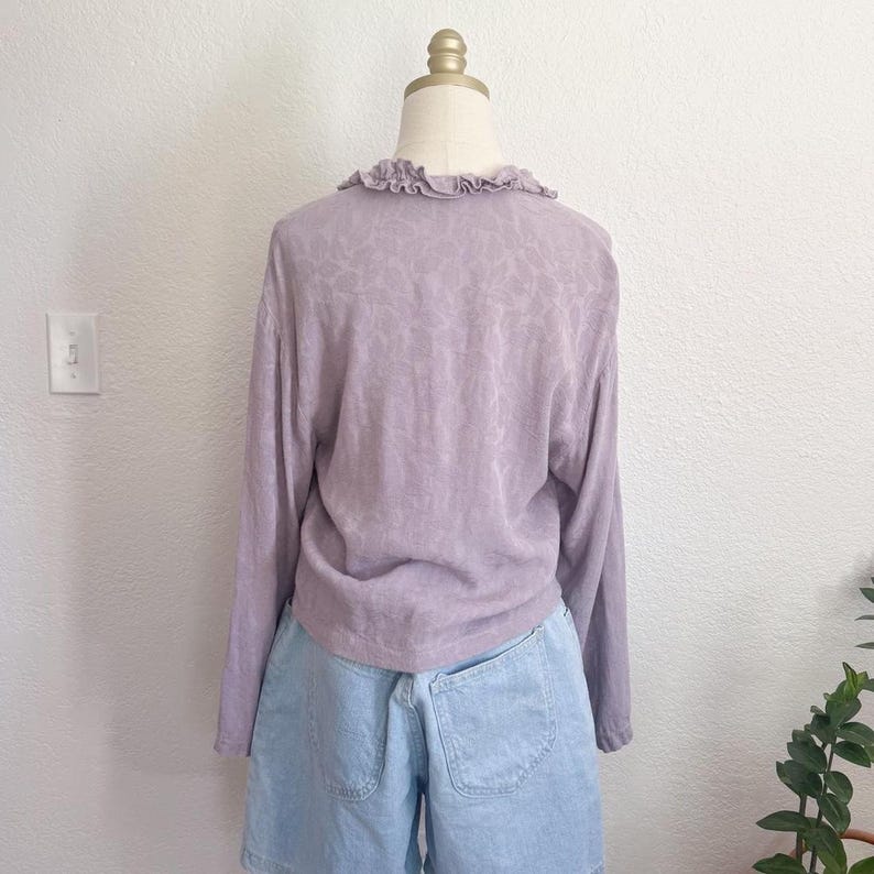 May include: A light purple long-sleeve shirt with a ruffled collar and a floral pattern. The shirt is paired with light blue denim shorts. The outfit is displayed on a mannequin against a white wall.