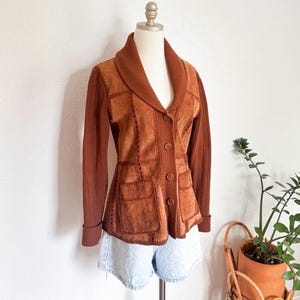May include: A brown suede jacket with a ribbed knit collar and sleeves. The jacket has a button-up front and two front pockets. The jacket is displayed on a mannequin with light blue denim shorts.