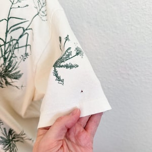 K&ouml;nnte beinhalten: Cremefarbenes T-Shirt mit einem dunkelgr&uuml;nen botanischen Druck. Das Design zeigt verschiedene Pflanzen und Gr&auml;ser. Der &Auml;rmel ist sichtbar und zeigt die detaillierte Illustration. Das Shirt besteht aus einem weichen, hellen Stoff.