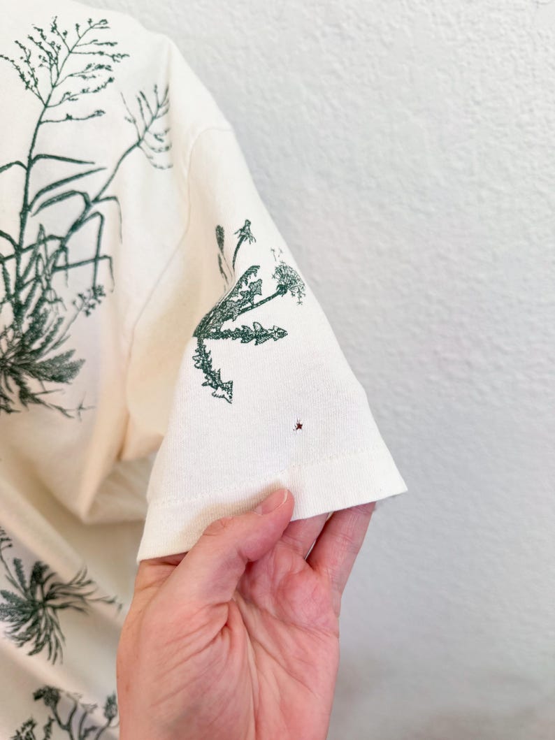 K&ouml;nnte beinhalten: Cremefarbenes T-Shirt mit einem dunkelgr&uuml;nen botanischen Druck. Das Design zeigt verschiedene Pflanzen und Gr&auml;ser. Der &Auml;rmel ist sichtbar und zeigt die detaillierte Illustration. Das Shirt besteht aus einem weichen, hellen Stoff.