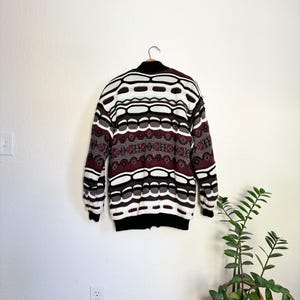 Peut inclure: Un pull &agrave; motif blanc, marron et bordeaux avec une fermeture &eacute;clair. Le pull est suspendu &agrave; un cintre contre un mur blanc.