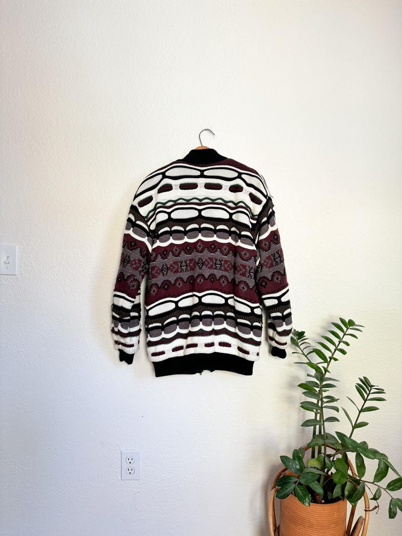 Peut inclure: Un pull &agrave; motif blanc, marron et bordeaux avec une fermeture &eacute;clair. Le pull est suspendu &agrave; un cintre contre un mur blanc.