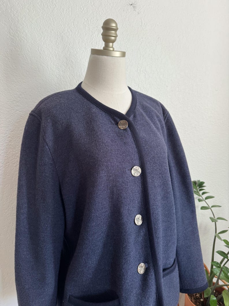 May include: A navy blue cardigan with a round neckline and dark blue trim. The cardigan has three silver-coloured buttons and two front pockets. The garment is displayed on a mannequin.