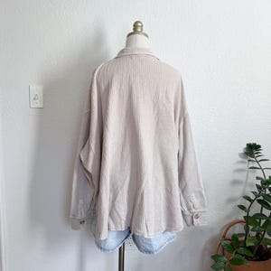 May include: Beige waffle-knit shirt with long sleeves and buttoned cuffs. The shirt is displayed on a mannequin, paired with light blue denim shorts. The shirt has a collared neckline and a relaxed fit, suitable for casual wear.