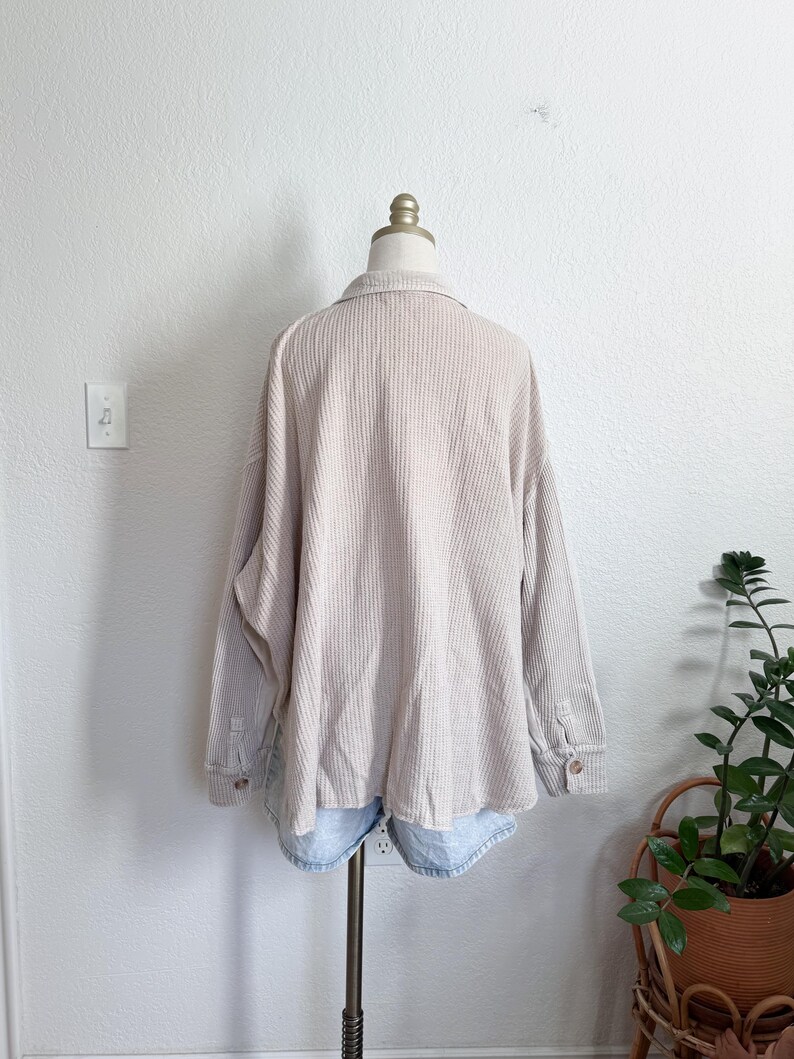 May include: Beige waffle-knit shirt with long sleeves and buttoned cuffs. The shirt is displayed on a mannequin, paired with light blue denim shorts. The shirt has a collared neckline and a relaxed fit, suitable for casual wear.