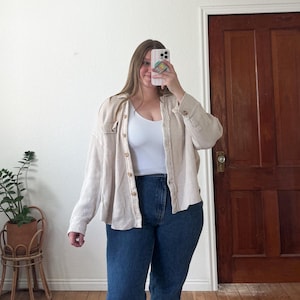 May include: A beige button-down shirt with a waffle texture is worn over a white ribbed tank top and dark blue jeans. The outfit is completed with a phone in hand, reflecting the image. The background includes a wooden door and a plant.