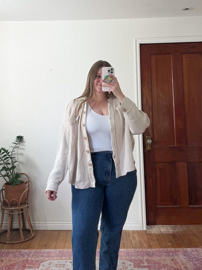 May include: A beige button-down shirt with a waffle texture is worn over a white ribbed tank top and dark blue jeans. The outfit is completed with a phone in hand, reflecting the image. The background includes a wooden door and a plant.