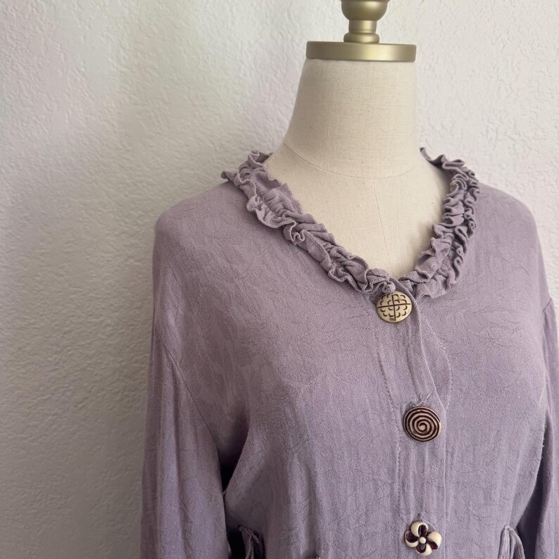May include: A lavender-colored blouse with a ruffled collar and decorative wooden buttons. The blouse has long sleeves and a textured fabric. The buttons have unique designs, including a spiral and a floral pattern. The blouse is displayed on a mannequin.