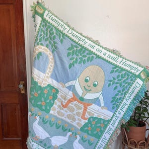May include: A woven throw blanket featuring the nursery rhyme character Humpty Dumpty. The blanket has a light blue background with Humpty Dumpty on a wall, geese, and the text "Humpty Dumpty sat on a wall" and "Horses and all the King's". The edges are trimmed with fringe.