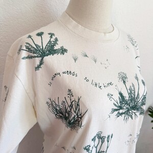 K&ouml;nnte beinhalten: Cremefarbenes T-Shirt mit Rundhalsausschnitt und botanischem Druck verschiedener Unkr&auml;uter in Gr&uuml;nt&ouml;nen. Der Text "so many weeds so little time" ist auf dem T-Shirt aufgedruckt. Das T-Shirt wird auf einer Schaufenquin pr&auml;sentiert.