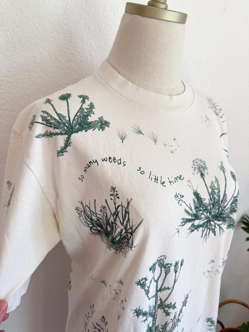 K&ouml;nnte beinhalten: Cremefarbenes T-Shirt mit Rundhalsausschnitt und botanischem Druck verschiedener Unkr&auml;uter in Gr&uuml;nt&ouml;nen. Der Text "so many weeds so little time" ist auf dem T-Shirt aufgedruckt. Das T-Shirt wird auf einer Schaufenquin pr&auml;sentiert.