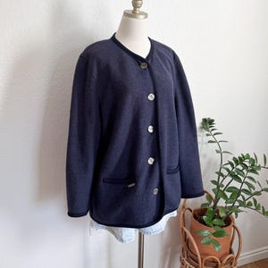 May include: A navy blue jacket with a round collar and silver buttons. The jacket has a dark blue trim and two front pockets. The jacket is displayed on a mannequin. A small plant in a wicker stand is in the background.
