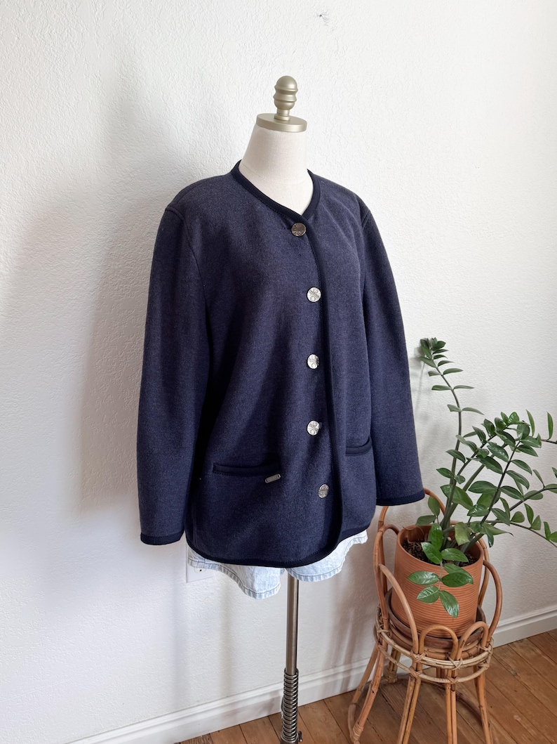 May include: A navy blue jacket with a round collar and silver buttons. The jacket has a dark blue trim and two front pockets. The jacket is displayed on a mannequin. A small plant in a wicker stand is in the background.