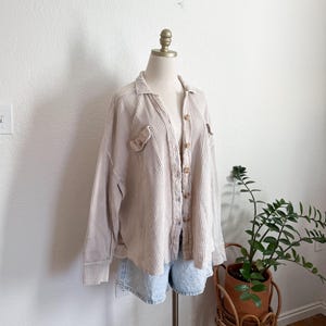 May include: Beige corduroy button-up shirt with two chest pockets and long sleeves. The shirt is displayed on a mannequin with light blue denim shorts. A potted plant in a wicker stand is in the background.