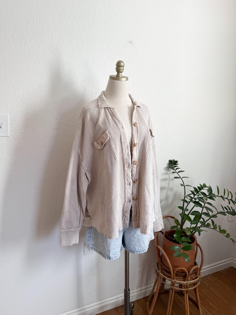 May include: Beige corduroy button-up shirt with two chest pockets and long sleeves. The shirt is displayed on a mannequin with light blue denim shorts. A potted plant in a wicker stand is in the background.