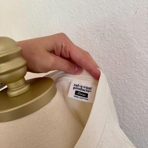 K&ouml;nnte beinhalten: Cremefarbenes T-Shirt mit einem Etikett mit der Aufschrift "rel-e-vant products" und "Large 100% Cotton". Das Shirt wird auf einer Schaufensterpuppe pr&auml;sentiert. Das Etikett ist wei&szlig; mit schwarzem Text.