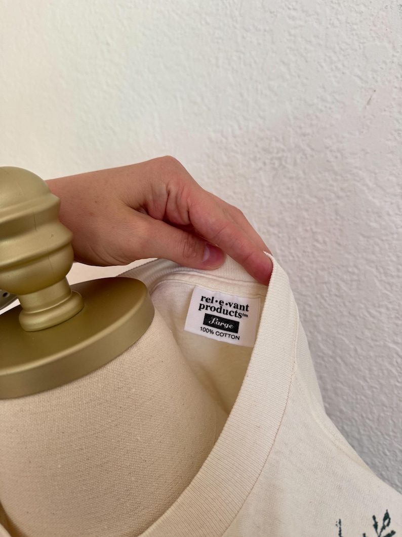 K&ouml;nnte beinhalten: Cremefarbenes T-Shirt mit einem Etikett mit der Aufschrift "rel-e-vant products" und "Large 100% Cotton". Das Shirt wird auf einer Schaufensterpuppe pr&auml;sentiert. Das Etikett ist wei&szlig; mit schwarzem Text.