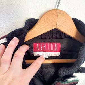 Peut inclure: Pullover ray&eacute; noir et blanc avec une &eacute;tiquette rouge et blanche indiquant "Ashton Cove" et une taille "M".