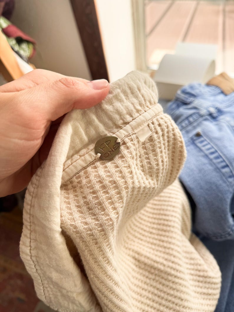 May include: Close-up of a beige waffle-knit fabric with a tag that reads "FP ONE". The fabric is part of a garment, possibly a top or pants, with a textured surface and a soft, light color.