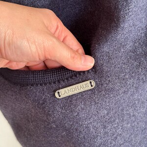 May include: A close-up shot of a navy blue woollen garment with a pocket. A small, rectangular silver tag with the word "LANDHAUS" is attached. The pocket is trimmed with a darker navy blue knitted fabric.
