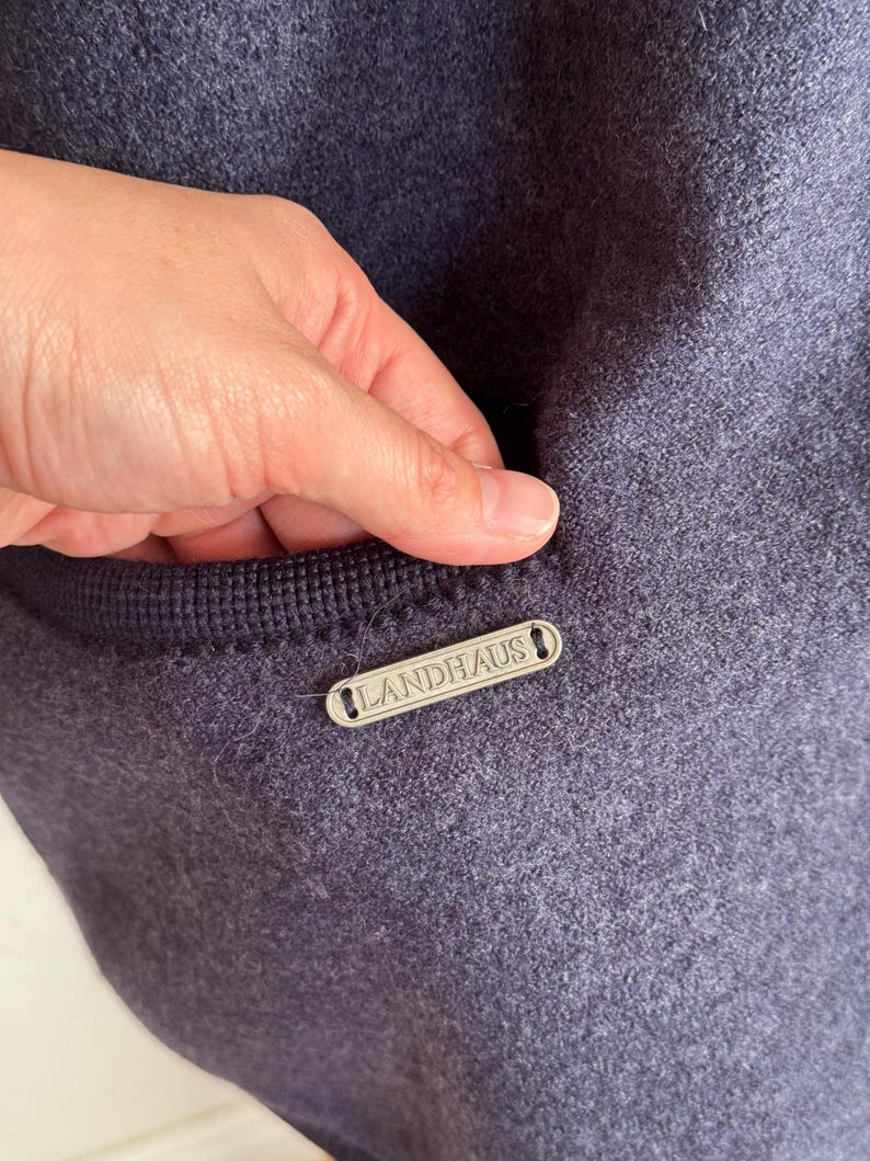 May include: A close-up shot of a navy blue woollen garment with a pocket. A small, rectangular silver tag with the word "LANDHAUS" is attached. The pocket is trimmed with a darker navy blue knitted fabric.