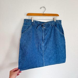 May include: A blue denim skirt with a belt loop and front pockets hangs on a wooden hanger. The skirt is a medium wash denim and appears to be vintage. The skirt is held by a person's hand.