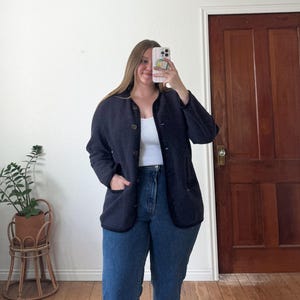 May include: A navy blue button-up jacket with black trim and two front pockets. The jacket is worn over a white vest top and paired with blue jeans. The outfit is casual and suitable for everyday wear.