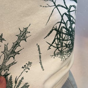 K&ouml;nnte beinhalten: Cremefarbener Stoff mit einem dunkelgr&uuml;nen botanischen Druck, der verschiedene Pflanzenillustrationen zeigt. Der Text "&copy;1994 rel-e-vant products" ist auf den Stoff gedruckt. Der Stoff ist leicht zerknittert.
