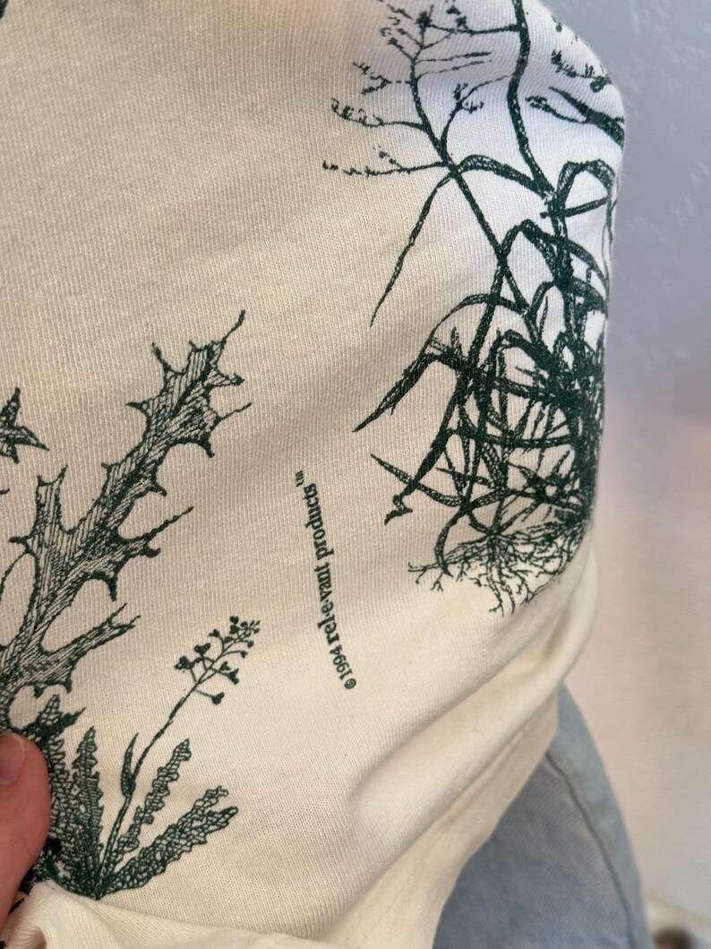 K&ouml;nnte beinhalten: Cremefarbener Stoff mit einem dunkelgr&uuml;nen botanischen Druck, der verschiedene Pflanzenillustrationen zeigt. Der Text "&copy;1994 rel-e-vant products" ist auf den Stoff gedruckt. Der Stoff ist leicht zerknittert.